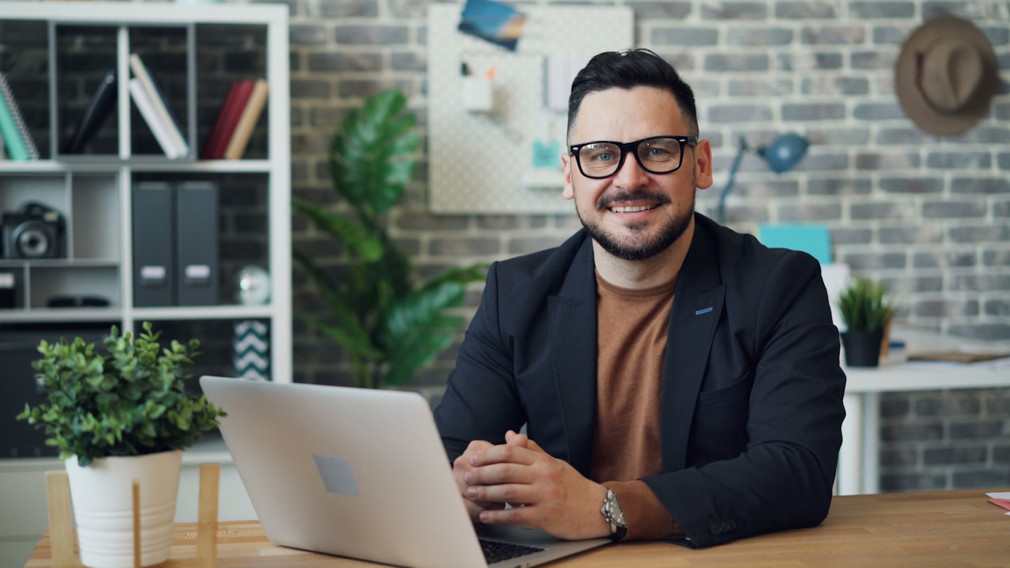 Mężczyzna z brodą i okularami uśmiecha się do laptopa na biurku.